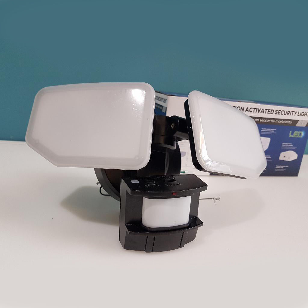 A dual-panel LED motion sensor light with a black adjustable mount and motion sensor base, shown on a white surface with its product box in the background against a teal wall.