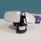 A dual-panel LED motion sensor light with a black adjustable mount and motion sensor base, shown on a white surface with its product box in the background against a teal wall.