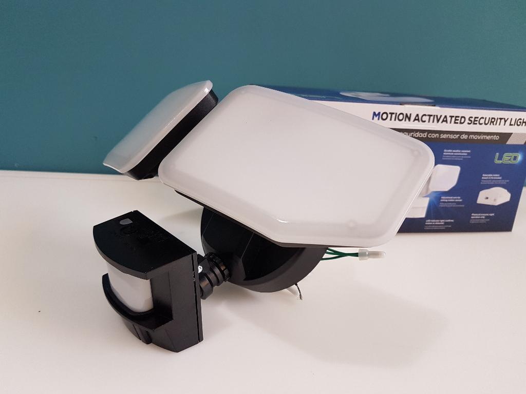 A motion-activated dual-slash security light with a white rectangular diffuser and black housing, shown alongside its blue and white product box on a white surface against a teal background.