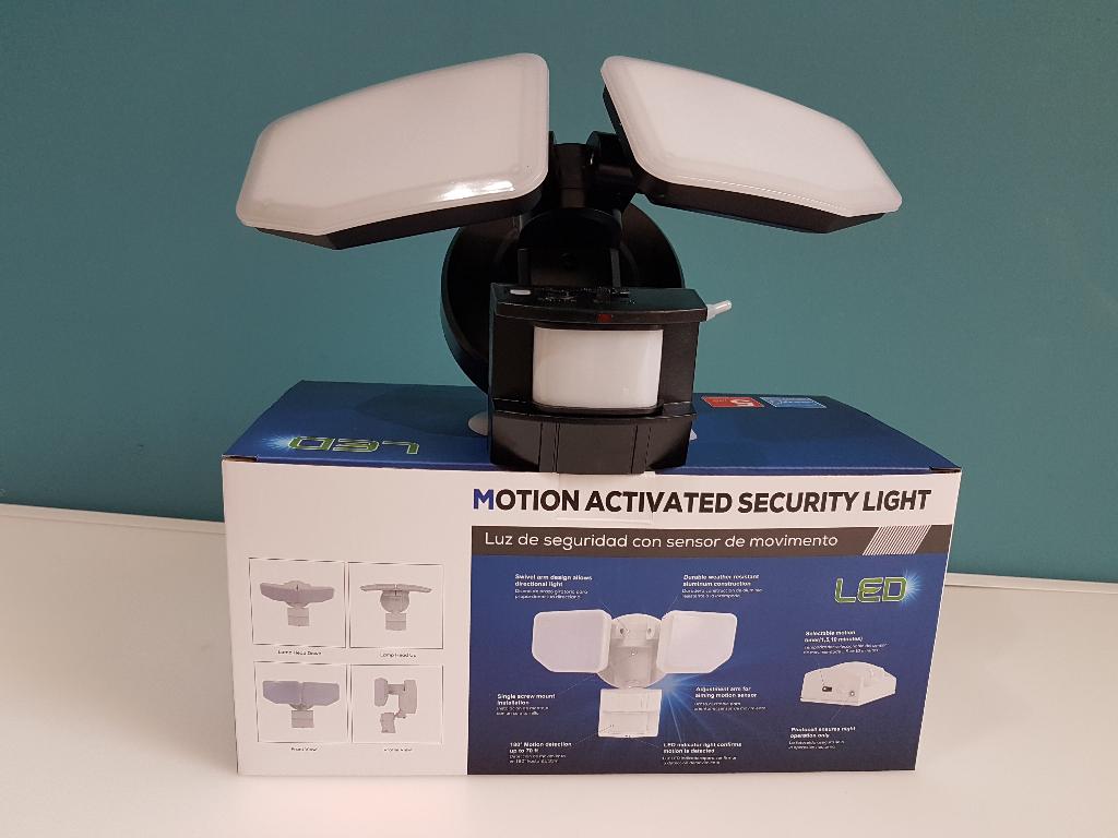 A motion-activated dual-slash LED security light with a black pole and white angled panels, displayed on top of its blue and white retail box against a teal wall.