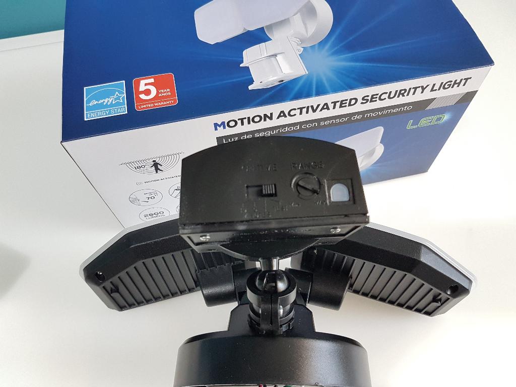 Black motion-activated security light with two angled solar panels, shown in front of its blue and white retail box.