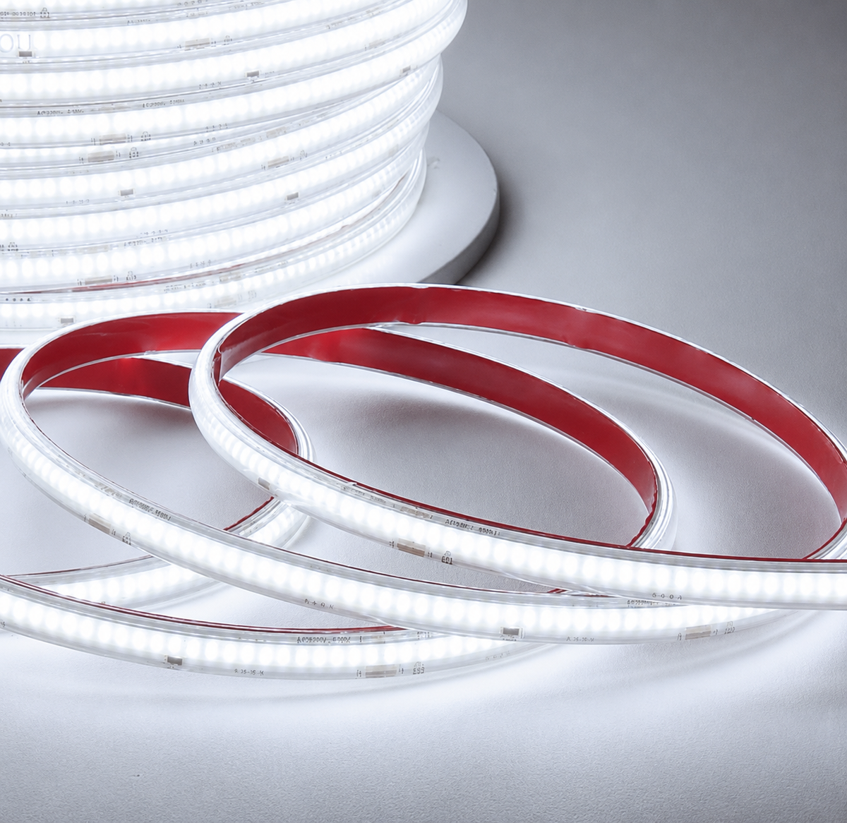 A coiled portion of high voltage COB LED light strip glowing with cool white light, shown in close-up against a warm beige background, next to the original spool of COB light strip.